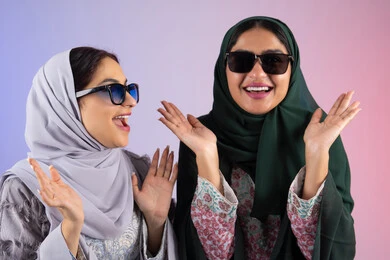 ارتداء النظارات الشمسيه للحمايه من اشعه الشمس ،بورتريه مقرب لسيدتان عربيتان خليجيتان اماراتيان ترتديان الحجاب والعباءة مبتسمتان ، تنظران بإيماءات الثقة بالنفس والفرح، مفهوم الأنوثة والنعومة، صديقتان سعوديتان تضعان ايديهم تحت وجههم بتعابير البهجه ، الاهتمام بالأناقة والمظهر الخارجي، ايماءات السرور والسعادة،  خلفية ملونه
