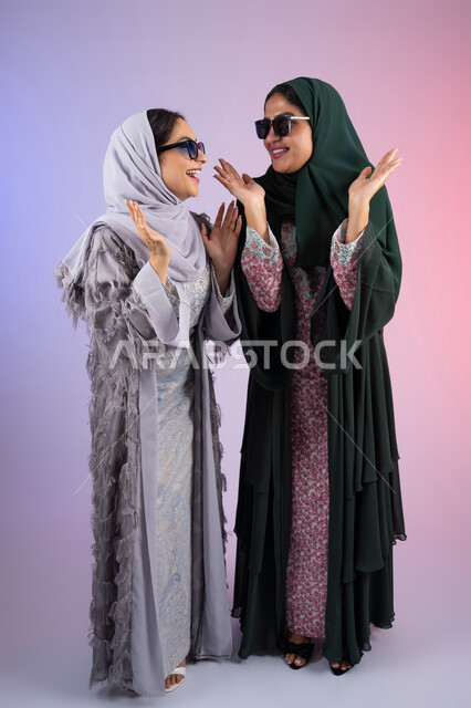 Looking at each other with gestures of pleasure and happiness, wearing sunglasses to protect from the sun's rays, a portrait of two Arab Gulf Emirati women wearing the hijab and abaya, smiling, looking with gestures of self-confidence and joy, the concept of femininity and softness, two Saudi friends putting their hands under their faces with expressions of joy, interest in elegance and external appearance, colorful background