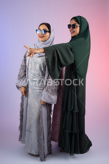 الاشاره الى شئ ما بايماءات البهجه والسعادة،  ارتداء النظارات الشمسيه للحمايه من اشعه الشمس ،بورتريه لسيدتان عربيتان خليجيتان اماراتيان ترتديان الحجاب والعباءة مبتسمتان ، تنظران بإيماءات الثقة بالنفس والفرح، مفهوم الأنوثة والنعومة، صديقتان سعوديتان تضعان ايديهم تحت وجههم بتعابير السرور، الاهتمام بالأناقة والمظهر الخارجي،  خلفية ملونه