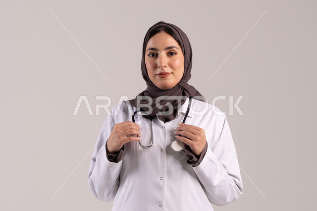 تقديم المساندة الطبية للمرضى, مهن ووظائف نسائية, , شابة سعودية تقوم بالعمل في المراكز والمستشفيات, بورتريه  لطبيبة عربية خليجية اماراتية ترتدي المعطف الطبي الأبيض وسماعة الفحص تنظر إلى الكاميرا بايماءات الثقة بالنفس, مفهوم الطب والرعاية الصحية, خلفية بنية