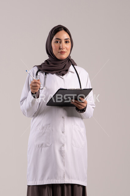 تسجيل البيانات والملاحظات المهمة, العمل في المراكز والمستشفيات، الطب والرعاية الصحية، بورتريه لطبيبة عربية خليجية اماراتية محجبة ترتدي المعطف الطبي الأبيض تضع سماعة الفحص الطبية تحمل بيدها حامل الاوراق, شابة سعودية تقوم بتقديم المساعدة الطبية، خلفية بنية