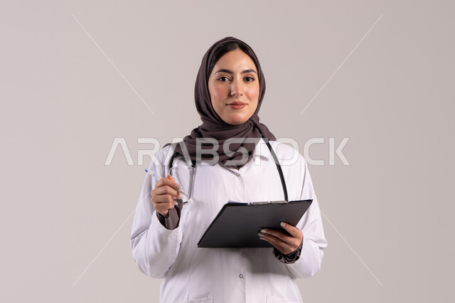كتابة البيانات والملاحظات اليومية, العمل في المراكز والمستشفيات، الطب والرعاية الصحية، بورتريه لطبيبة عربية خليجية اماراتية محجبة ترتدي المعطف الطبي الأبيض تضع سماعة الفحص الطبية تحمل بيدها حامل الاوراق, شابة سعودية تقوم بتقديم المساعدة الطبية، خلفية بنية