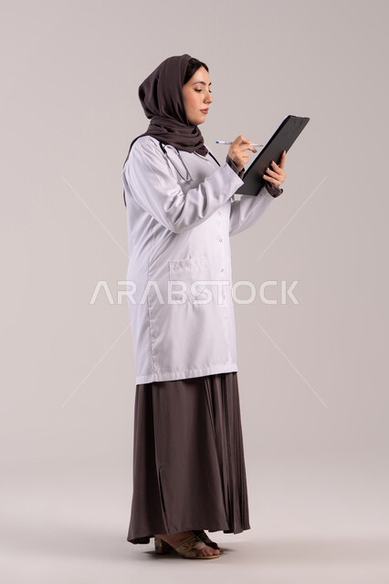Recording notes and information, working in centers and hospitals, medicine and healthcare, a portrait of an Arab Gulf Emirati female doctor wearing a white medical coat, holding a stethoscope and a clipboard in her hand, a young Saudi woman providing medical assistance, full-body, brown background.