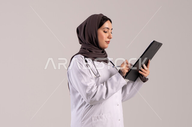 العمل المهني بالمستشفيات والمراكز الطبية، الطب والرعاية الصحية، تدوين الملاحظات والبيانات، بورتريه من الجانب لطبيبة عربية خليجية اماراتية محجبة ترتدي المعطف الطبي الأبيض تضع سماعة الفحص الطبية تحمل بيدها حامل الاوراق, شابة سعودية تقوم بتقديم المساعدة الطبية، خلفية بنية