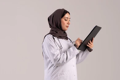 العمل المهني بالمستشفيات والمراكز الطبية، الطب والرعاية الصحية، تدوين الملاحظات والبيانات، بورتريه من الجانب لطبيبة عربية خليجية اماراتية محجبة ترتدي المعطف الطبي الأبيض تضع سماعة الفحص الطبية تحمل بيدها حامل الاوراق, شابة سعودية تقوم بتقديم المساعدة الطبية، خلفية بنية