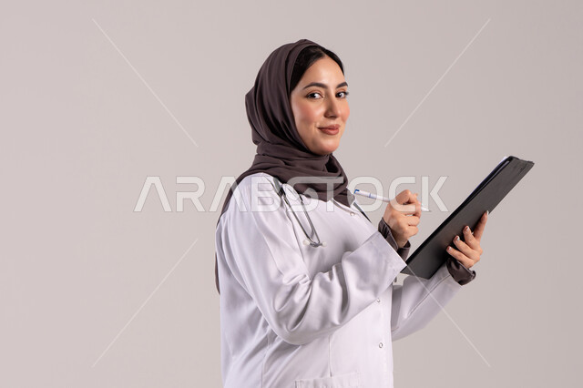 شابة سعودية تقوم بتقديم المساعدة الطبية, العمل في المراكز والمستشفيات، الطب والرعاية الصحية، تدوين الملاحظات والبيانات، بورتريه من الجانب لطبيبة عربية خليجية اماراتية محجبة ترتدي المعطف الطبي الأبيض تضع سماعة الفحص الطبية تحمل بيدها حامل الاوراق، خلفية بنية