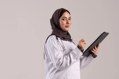 شابة سعودية تقوم بتقديم المساعدة الطبية, العمل في المراكز والمستشفيات، الطب والرعاية الصحية، تدوين الملاحظات والبيانات، بورتريه من الجانب لطبيبة عربية خليجية اماراتية محجبة ترتدي المعطف الطبي الأبيض تضع سماعة الفحص الطبية تحمل بيدها حامل الاوراق، خلفية بنية