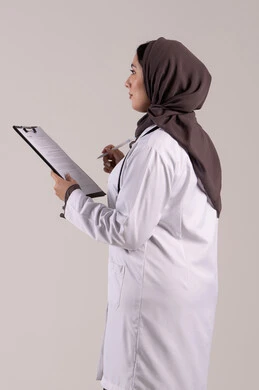 الطب والرعاية الصحية, العمل في المراكز والمستشفيات، تدوين الملاحظات والبيانات، بورتريه من الجانب لطبيبة عربية خليجية اماراتية محجبة ترتدي المعطف الطبي الأبيض تضع سماعة الفحص الطبية تحمل بيدها حامل الاوراق, شابة سعودية تقوم بتقديم المساعدة الطبية، خلفية بنية