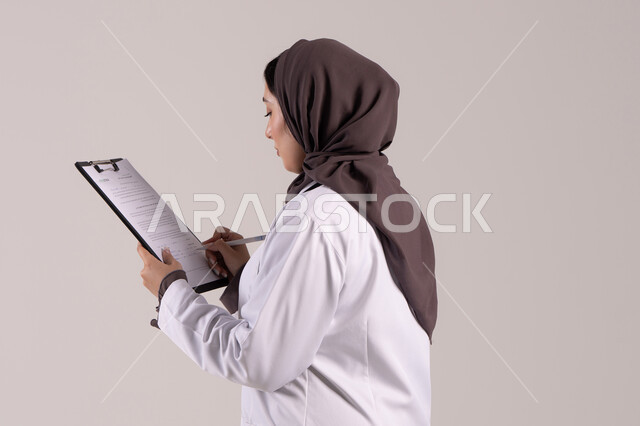 العمل في المراكز والمستشفيات، الطب والرعاية الصحية، تدوين الملاحظات والبيانات، بورتريه مقرب من الجانب لطبيبة عربية خليجية اماراتية محجبة ترتدي المعطف الطبي الأبيض تضع سماعة الفحص الطبية تحمل بيدها حامل الاوراق, شابة سعودية تقوم بتقديم المساعدة الطبية، خلفية بنية