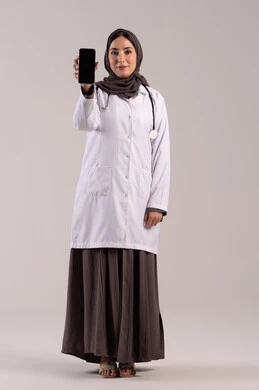A blank black screen, displaying health applications and programs, a portrait of an Arab Gulf Emirati female doctor wearing a white coat and stethoscope, holding a mobile phone in her hand and looking at the camera, using modern technological devices and integrating technology with medicine, full-body shot, brown background.