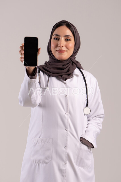 عرض التطبيقات والبرامج الصحية، بورتريه لطبيبة عربيه خليجيه اماراتية ترتدي المعطف الأبيض و السماعة تحمل بيدها الهاتف المحمول تنظر إلى الكاميرا، استخدام الأجهزة التقنية الحديثة ودمج التكنولوجيا مع الطب، عرض شاشة سوادء فارغة، خلفية بنية