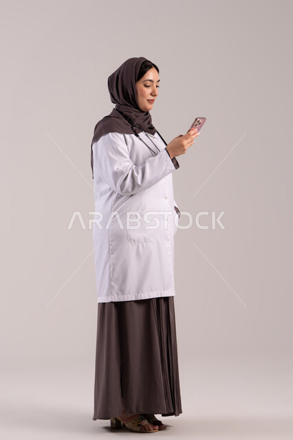 E-health programs, providing services and healthcare online, a side portrait of an Arab Gulf Emirati female doctor wearing a medical coat and stethoscope using a mobile phone, utilizing modern technological devices and integrating technology with medicine, full-body shot, brown background.