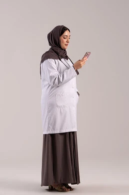 برامج الصحة الإلكترونية, تقديم الخدمات والرعاية الصحية عبر الإنترنت، بورتريه من الجانب لطبيبة عربيه خليجيه امارتية ترتدي المعطف الطبي و السماعه تقوم باستخدام الهاتف المحمول، استخدام الأجهزة التقنية الحديثة ودمج التكنولوجيا مع الطب الجسم بالطول الكامل، خلفية بنية