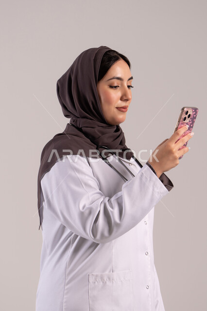 التطبيقات الرقمية للخدمات الصحية, تقديم الخدمات والرعاية الصحية عبر الإنترنت، بورتريه من الجانب لطبيبة عربيه خليجيه امارتية ترتدي المعطف الطبي و السماعه تقوم باستخدام الهاتف المحمول، استخدام الأجهزة التقنية الحديثة ودمج التكنولوجيا مع الطب، خلفية بنية