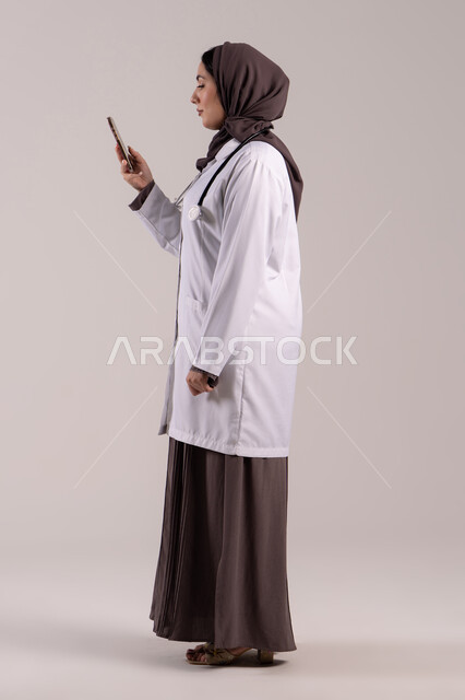 البرامج والتطبيقات الخاصة بالصحة, تقديم الخدمات والرعاية الصحية عبر الإنترنت، بورتريه من الجانب لطبيبة عربيه خليجيه امارتية ترتدي المعطف الطبي و السماعه تقوم باستخدام الهاتف المحمول، استخدام الأجهزة التقنية الحديثة ودمج التكنولوجيا مع الطب، الجسم بالطول الكامل, خلفية بنية