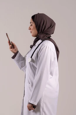 Health applications and programs, providing online health services and care, a side portrait of an Arab Gulf Emirati female doctor wearing a medical coat and stethoscope using a mobile phone, utilizing modern technological devices and integrating technology with medicine, brown background.