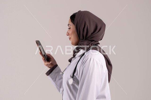 استخدام الأجهزة التقنية الحديثة ودمج التكنولوجيا مع الطب, تقديم الخدمات والرعاية الصحية عبر الإنترنت، بورتريه من الجانب لطبيبة عربيه خليجيه امارتية ترتدي المعطف الطبي و السماعه تقوم باستخدام الهاتف المحمول، التطبيقات والبرامج الصحية، خلفية بنية