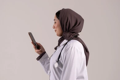 The use of modern technical devices and the integration of technology with medicine, providing online health services and care, a side portrait of an Arab Gulf Emirati female doctor wearing a medical coat and stethoscope using a mobile phone, health applications and programs, brown background.