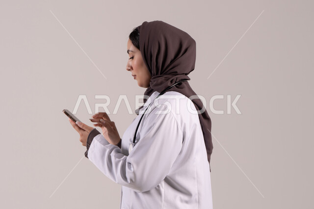 تقديم الخدمات والرعاية الصحية عبر الإنترنت، بورتريه من الجانب لطبيبة عربيه خليجيه امارتية ترتدي المعطف الطبي و السماعه تقوم باستخدام الهاتف المحمول، استخدام الأجهزة التقنية الحديثة ودمج التكنولوجيا مع الطب، التطبيقات والبرامج الصحية، خلفية بنية