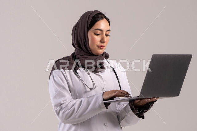 تسجيل وتدوين معلومات وملفات المرضى عبر الابتوب, دمج التقنية الحديثة في مجال العمل الطبي, بورترية لطبيبة عربية خليجية اماراتية ترتدي المعطف الطبي الأبيض وسماعة الفحص تحمل الحاسب المحمول, شابة سعودية تقوم بتقديم الخدمات العلاجية في المراكز الطبية والمستشفيات , خلفية بنيه