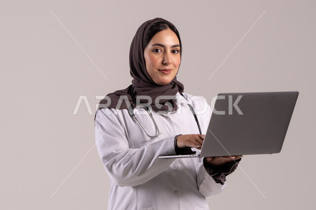 إدارة ملفات المرضى إلكترونيًا على اللابتوب, دمج التقنية الحديثة في مجال العمل الطبي, بورترية لطبيبة عربية خليجية اماراتية ترتدي المعطف الطبي الأبيض وسماعة الفحص تحمل الحاسب المحمول, شابة سعودية تقوم بتقديم الخدمات العلاجية في المراكز الطبية والمستشفيات , خلفية بنيه