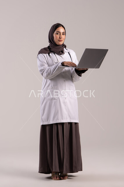 A young Saudi woman provides therapeutic services in medical centers and hospitals, recording and documenting patient information and files via a laptop, integrating modern technology in the field of medical work, a portrait of an Arab Gulf Emirati doctor wearing a white medical coat and a stethoscope holding a laptop, full-body, brown background.