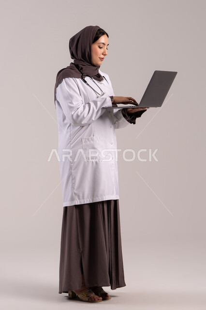دمج التقنية الحديثة في مجال العمل الطبي, تسجيل وتدوين معلومات وملفات المرضى عبر الابتوب, بورتريةمن الجانب لطبيبة عربية خليجية اماراتية ترتدي المعطف الطبي الأبيض وسماعة الفحص تحمل الحاسب المحمول, شابة سعودية تقوم بتقديم الخدمات العلاجية في المراكز الطبية والمستشفيات , الجسم بالطول الكامل, خلفية بنيه