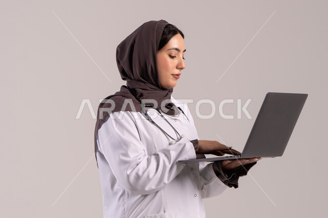 تسجيل وتدوين معلومات وملفات المرضى عبر الابتوب, دمج التقنية الحديثة في مجال العمل الطبي, بورترية لطبيبة عربية خليجية اماراتية ترتدي المعطف الطبي الأبيض وسماعة الفحص تحمل الحاسب المحمول, شابة سعودية تقوم بتقديم الخدمات العلاجية في المراكز الطبية والمستشفيات , خلفية بنيه