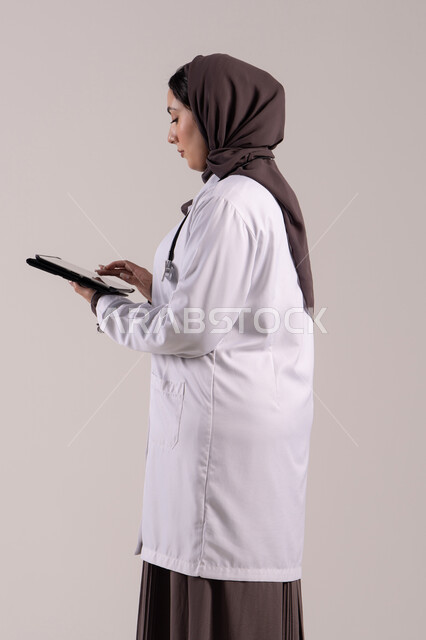 العمل في القطاع الصحي , تطور القطاع الصحي في الامارات العربية،  سيده سعودية تقوم بتقديم الاستشارات للمرضى عن بعد الأجهزة التكنولوجية، بورتريه لطبيبة عربية خليجية اماراتية ترتدي المعطف الأبيض و السماعة تمسك جهاز التابلت ، الجسم بالطول الكامل, خلفية بنية
