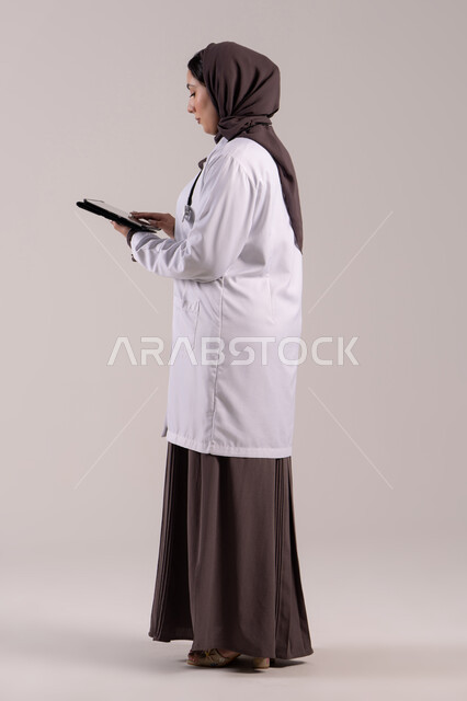 مفهوم الخدمات الطبية, تطور القطاع الصحي في الامارات العربية،  سيده سعودية تقوم بتقديم الاستشارات للمرضى عن بعد الأجهزة التكنولوجية، بورتريه من الجانب لطبيبة عربية خليجية اماراتية ترتدي المعطف الأبيض و السماعة تمسك جهاز التابلت ، الجسم بالطول الكامل, خلفية بنية