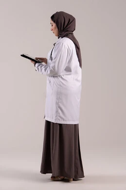 The concept of medical services, the development of the health sector in the United Arab Emirates, a Saudi woman providing remote consultations to patients using technological devices, a side portrait of an Emirati Gulf Arab female doctor wearing a white coat and stethoscope holding a tablet device, full body, brown background.