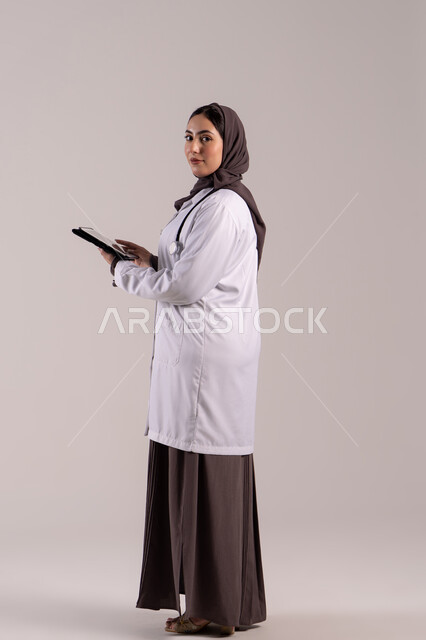 دمج التقنيات الحديثة بالمجالات الطبية, تطور القطاع الصحي في الامارات العربية،  سيده سعودية تقوم بتقديم الاستشارات للمرضى عن بعد الأجهزة التكنولوجية، بورتريه من الجانب لطبيبة عربية خليجية اماراتية ترتدي المعطف الأبيض و السماعة تمسك جهاز التابلت ، الجسم بالطول الكامل, خلفية بنية