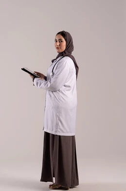 دمج التقنيات الحديثة بالمجالات الطبية, تطور القطاع الصحي في الامارات العربية،  سيده سعودية تقوم بتقديم الاستشارات للمرضى عن بعد الأجهزة التكنولوجية، بورتريه من الجانب لطبيبة عربية خليجية اماراتية ترتدي المعطف الأبيض و السماعة تمسك جهاز التابلت ، الجسم بالطول الكامل, خلفية بنية
