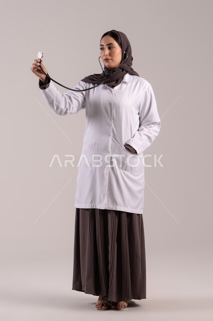 مفهوم الطب و الرعاية، بورتريه لطبيبة عربية خليجية اماراتية محجبة ترتدي المعطف الطبي وتحمل بيدها سماعة الطبيب لسماع نبضات القلب, سيده سعودية تعمل في مجال الرعاية الصحية وتقديم الخدمات الطبية المتميزة، الجسم بالطول الكامل, خلفية بنية