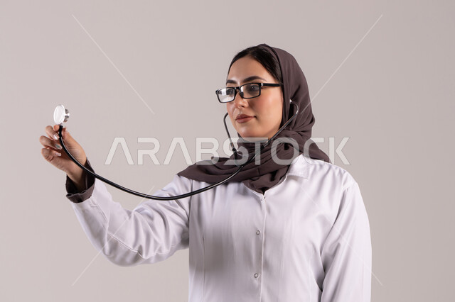 Working in the healthcare sector, a close-up portrait of an Emirati Arab female doctor wearing a medical coat and glasses, holding a stethoscope in her hand to listen to heartbeats. A Saudi woman working in the field of healthcare and providing distinguished medical services, developing the healthcare sector in the United Arab Emirates, with a brown background.