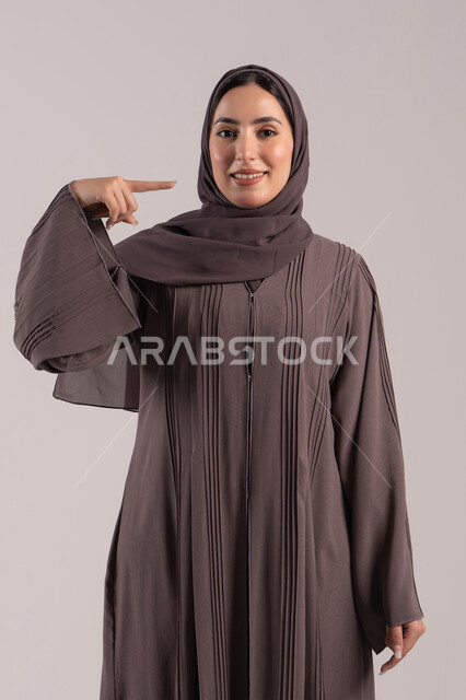 Using hand gestures to indicate the location here, identifying a specific object, a portrait of a veiled Emirati Gulf Arab woman wearing an abaya pointing with her index finger to her face, an elegant young Saudi woman, brown background.