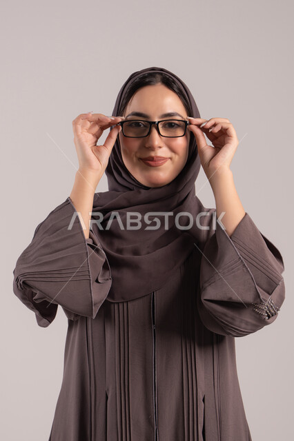 تحسين النظر وتوضيح الرؤية, الاناقة و الاهتمام بالمظهر الخارجي، بورتريه لامرأة عربية خليجية اماراتية محجبة ترتدي عباءة تمسك بيدها نظارة طبية، ارتداء النظارات الخاصة بضعف النظر، شابة سعودية انيقة، خلفية بنية