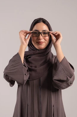 تحسين النظر وتوضيح الرؤية, الاناقة و الاهتمام بالمظهر الخارجي، بورتريه لامرأة عربية خليجية اماراتية محجبة ترتدي عباءة تمسك بيدها نظارة طبية، ارتداء النظارات الخاصة بضعف النظر، شابة سعودية انيقة، خلفية بنية