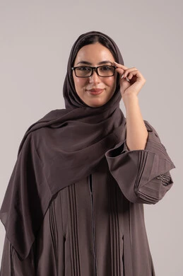 An elegant young Saudi woman, concerned about her appearance, a portrait of a veiled Emirati Gulf Arab woman wearing an abaya holding a pair of glasses in her hand, wearing glasses for vision impairment, improving vision and trying to correct eyesight, brown background.