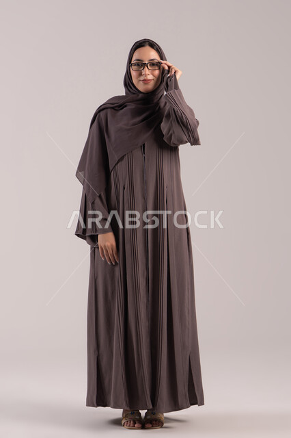تحسين الرؤية ومحاولة تعديل البصر,الاهتمام بالمظهر الخارجي، بورتريه لامرأة عربية خليجية اماراتية محجبة ترتدي عباءة تمسك بيدها نظارة طبية، ارتداء النظارات الخاصة بضعف النظر، شابة سعودية انيقة، الجسم بالطول الكامل ,خلفية بنية