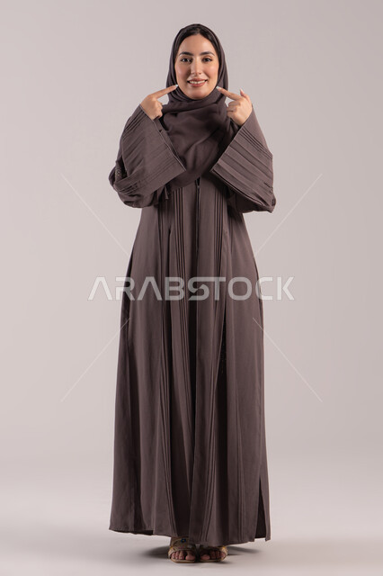 Identifying the location of a specific object, gestures and hand movements indicating this place (here), a portrait of a veiled Emirati Gulf Arab woman wearing an abaya pointing with her index finger to her face, an elegant young Saudi woman, full-body, brown background.