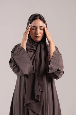 المعاناة من آلام الشقيقة, الحاجة للهدوء والراحة، الإحساس بألم الرأس والصداع، بورتريه لإمرأة عربية خليجية اماراتية محجبة ترتدي عباءة تضع يديها على رأسها بايماءات التعب والارهاق، شابة سعودية مريضة، خلفية بنية