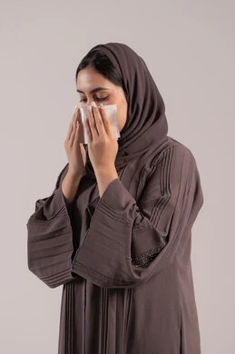 استخدام المناديل لحماية الصحة أثناء العطس، الشعور بالتعب والمرض، الإصابة بفايروس كورونا كوفيد19، بورتريه لامرأه عربيه خليجيه اماراتية محجبة ترتدي عباءة ، إيماءات وجه ويدين تدل على العطاس، شابة سعودية مريضة، خلفية بنية