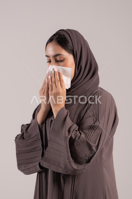 تغطية الفم بالمناديل عند العطس، الشعور بالتعب والمرض، الإصابة بفايروس كورونا كوفيد19، بورتريه لامرأه عربيه خليجيه اماراتية محجبة ترتدي عباءة ، إيماءات وجه ويدين تدل على العطاس، شابة سعودية مريضة، خلفية بنية