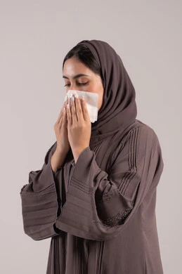 Covering the mouth with tissues when sneezing, feeling tired and sick, contracting the coronavirus COVID-19, a portrait of an Arab Gulf Emirati woman wearing a hijab and an abaya, facial and hand gestures indicating sneezing, a sick young Saudi woman, brown background.