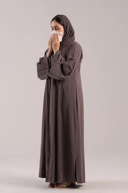 Infection with the coronavirus COVID-19, using tissue paper while sneezing, feeling tired and sick, a side portrait of an Arab Gulf Emirati woman wearing a hijab and an abaya, facial and hand gestures indicating sneezing, a sick young Saudi woman, full-body, brown background.