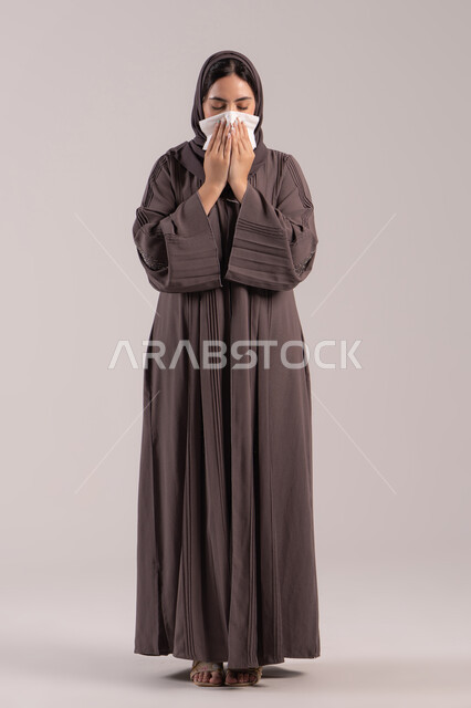 الشعور بالتعب والمرض, استعمال المناديل الورقية أثناء العطس، الإصابة بفايروس كورونا كوفيد19، بورتريه لامرأه عربيه خليجيه اماراتية محجبة ترتدي عباءة ، إيماءات وجه ويدين تدل على العطاس، شابة سعودية مريضة، الجسم بالطول الكامل, خلفية بنية