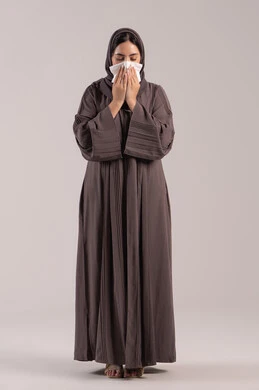 الشعور بالتعب والمرض, استعمال المناديل الورقية أثناء العطس، الإصابة بفايروس كورونا كوفيد19، بورتريه لامرأه عربيه خليجيه اماراتية محجبة ترتدي عباءة ، إيماءات وجه ويدين تدل على العطاس، شابة سعودية مريضة، الجسم بالطول الكامل, خلفية بنية