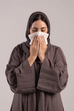 استعمال المناديل الورقية أثناء العطس، الشعور بالتعب والمرض، الإصابة بفايروس كورونا كوفيد19، بورتريه لامرأه عربيه خليجيه اماراتية محجبة ترتدي عباءة ، إيماءات وجه ويدين تدل على العطاس، شابة سعودية مريضة، خلفية بنية