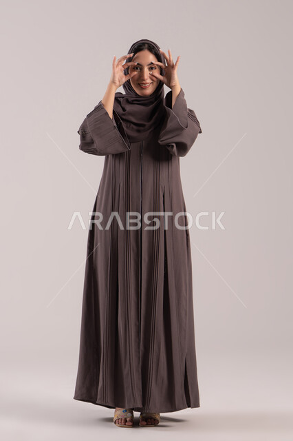 The concept of a future vision, a love for research, exploration, and adventure, a portrait of a smiling Emirati Gulf Arab woman in a hijab wearing a black abaya trying to bring distant things closer, a Saudi woman placing her hands over her eyes like a lens, full-body. Brown background.
